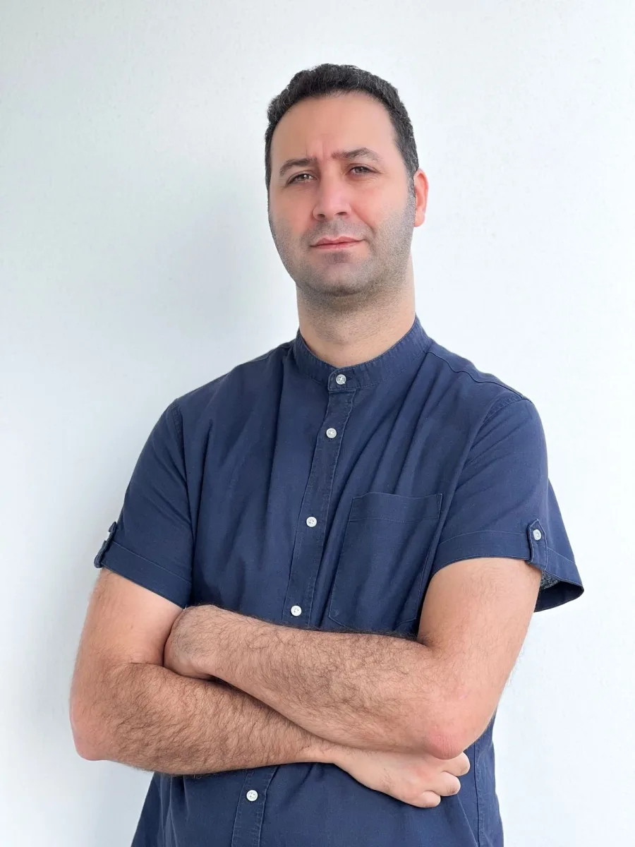 Koray Yalçın, beyaz duvar önünde lacivert gömlekle kollarını kavuşturmuş